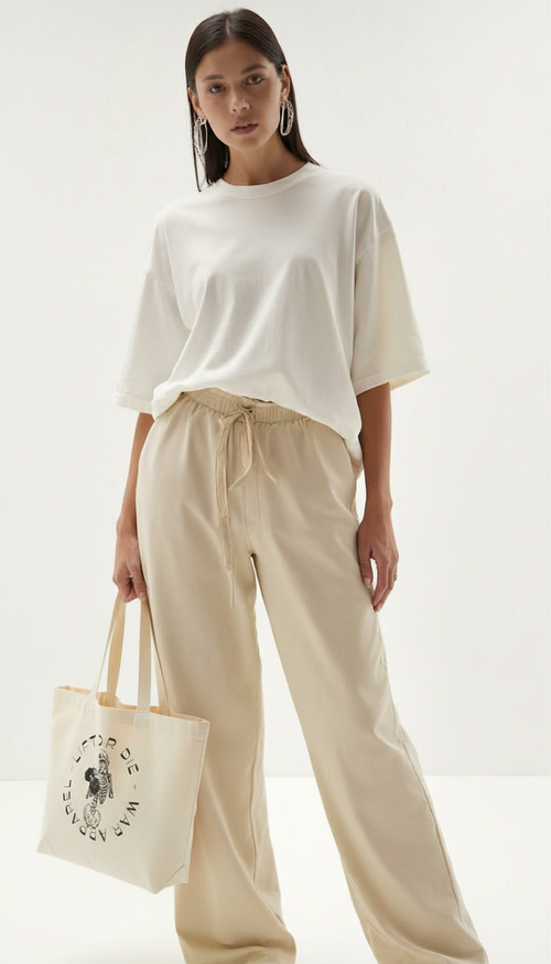 Woman holding a WAR APPAREL beige tote bag wearing a white top and beige pants on a white background